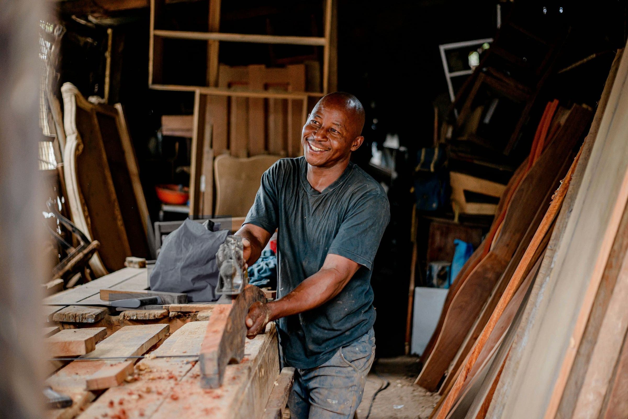 A carpenter smiles as he planes a length of timber at his workshop bench.