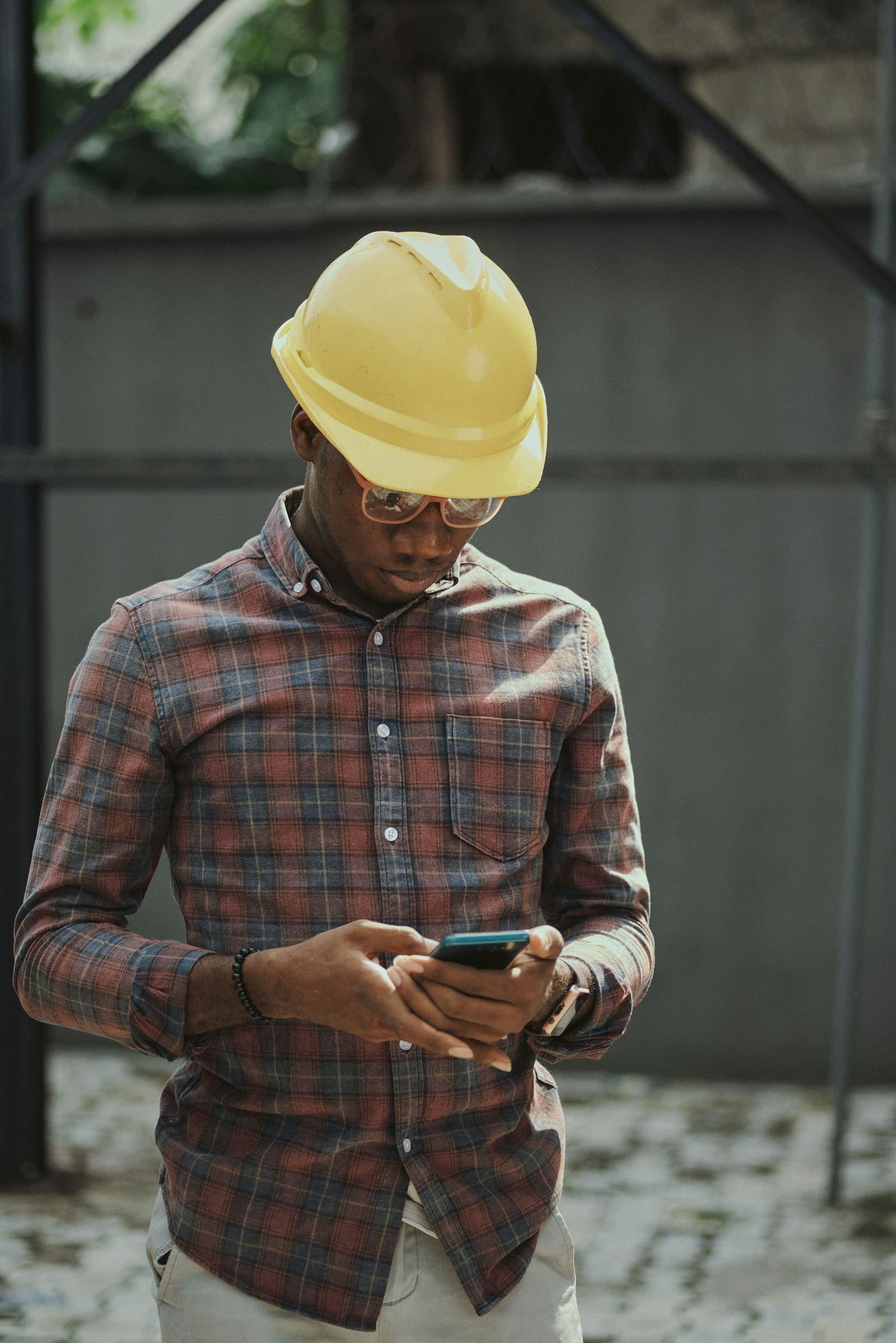 A site engineer in a yellow hard hat and safety glasses checks his phone outdoors.