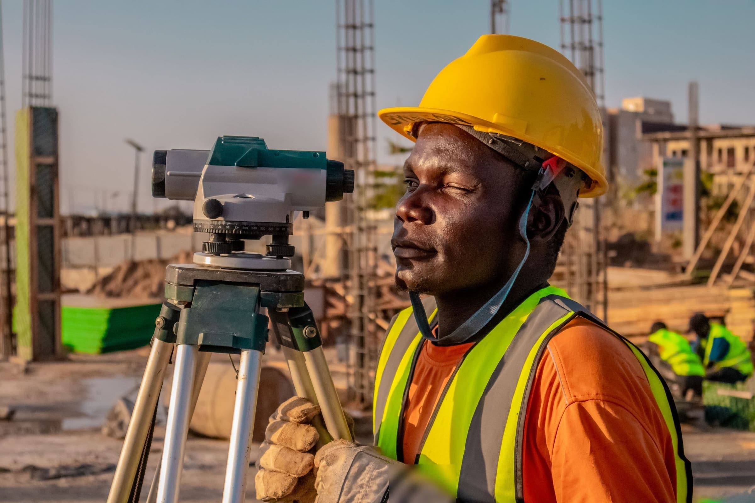 A surveyor in a hard hat and high-visibility vest beside an optical level on a construction site at sunset.