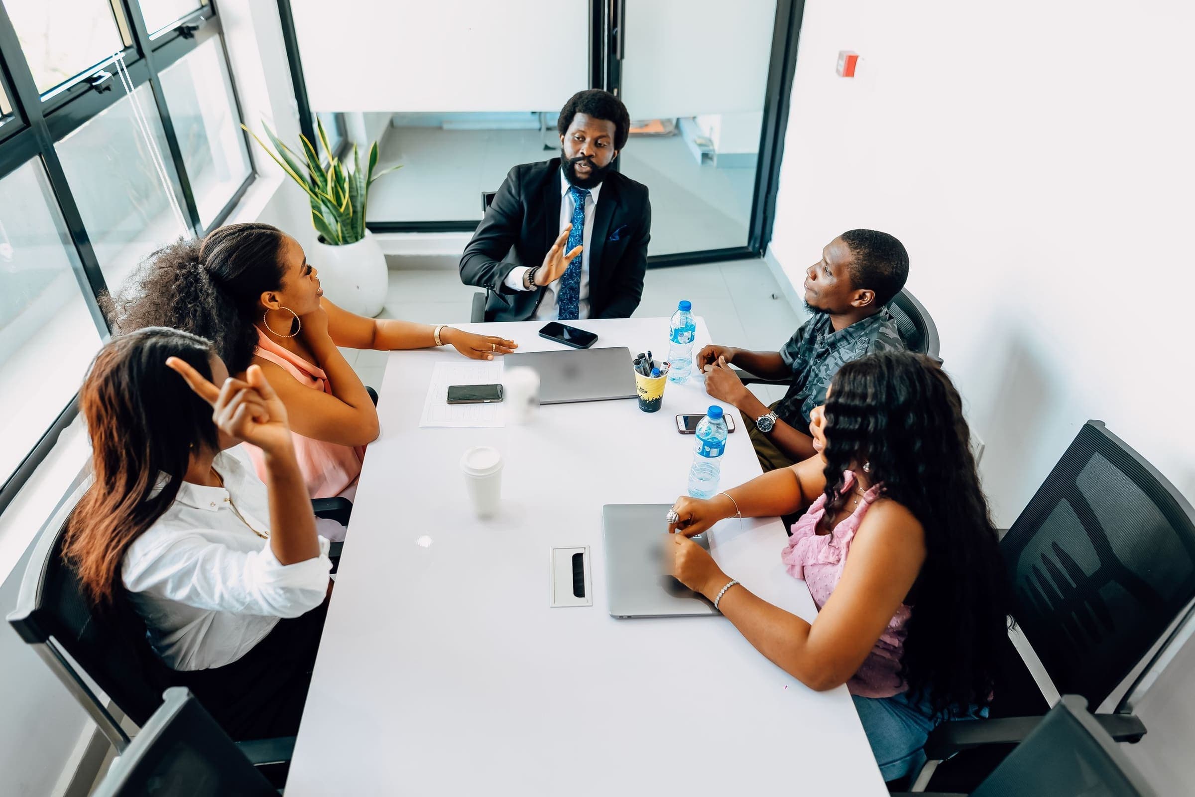 A leader in a dark suit leads a discussion with four colleagues around a meeting table in a bright office.